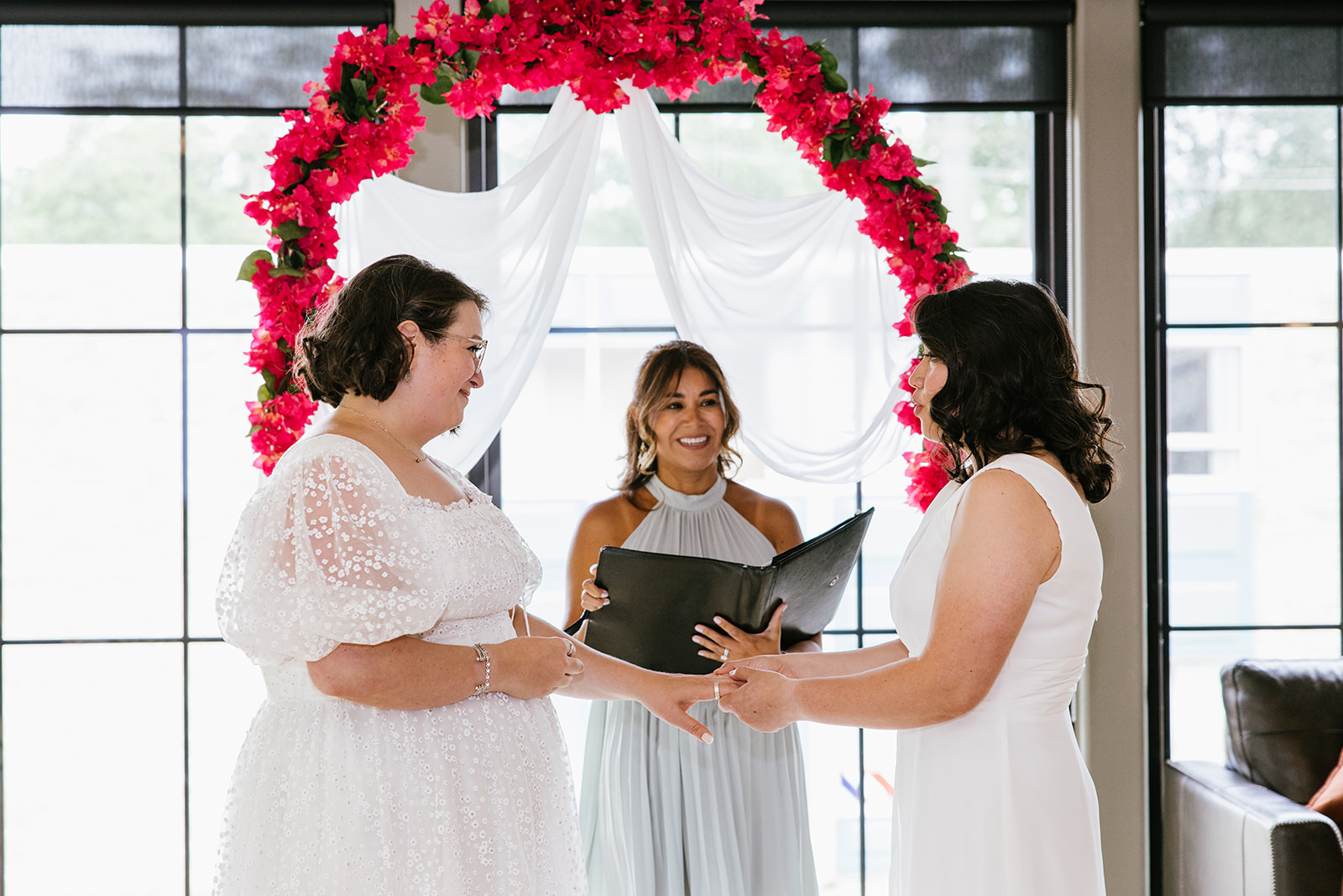 Two brides holding hands during a ceremony with bright pink flowers and white draping behind them