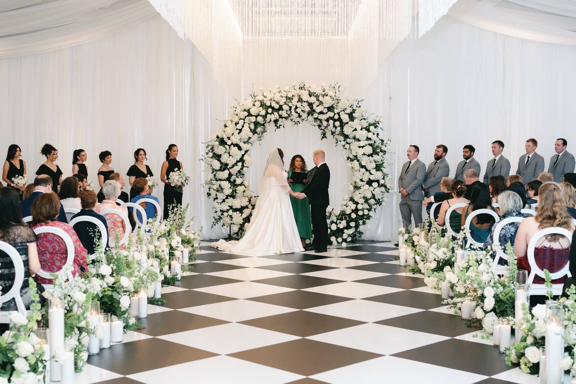 Elegant indoor wedding ceremony beneath a grand floral arch with guests seated along a black-and-white checkered aisle