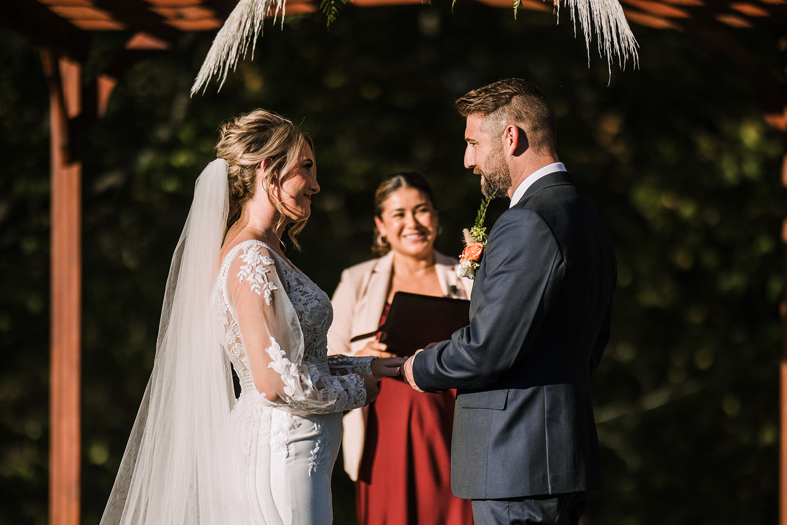 Bride and groom exchanging vows outdoors beneath a wooden pergola with officiant smiling in the background